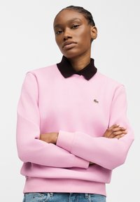 Νεαρό άτομο με πλεξούδες, φορώντας ένα ροζ φούτερ με μαύρο γιακά και μπλε τζιν, στέκεται με σταυρωμένα τα χέρια.