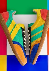 Paire de baskets en daim colorées avec des panneaux jaunes, verts, bleus, orange, rouges et noirs sur un fond multicolore.