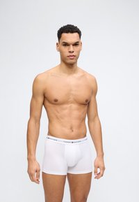 Oberkörperfreier Mann in weißen Tommy Hilfiger Boxershorts, der vor einem einfarbigen hellen Hintergrund steht und mit neutralem Gesichtsausdruck geradeaus blickt.