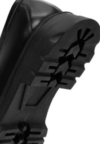 Chaussure en cuir noir avec une semelle en caoutchouc texturée, dotée de rainures profondes pour l'adhérence et d'une conception de bout solide et arrondi.