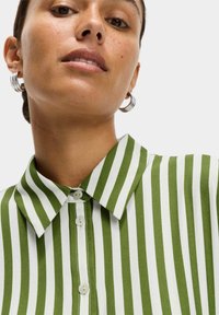 Camicia a righe verdi e bianche con collo, dotata di una chiusura a bottoni frontale, caratterizzata da una texture liscia e chiusura a due bottoni sul collo.