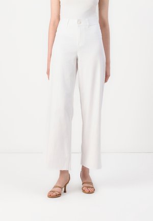 Kvinde iført hvide, højtaljede bukser med vide ben og rå kanter samt beige åbne hæle, stående foran en hvid baggrund.