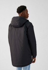 Chaqueta negra con capucha de diseño largo, puños elásticos y cuello con marca, combinada con jeans de un azul claro. Textura de tela suave.