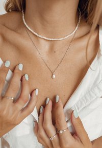 Drie kettingen zijn gestapeld: een witte kralen chokers, een zilveren ketting en een hanger met een druppelvormige parel.