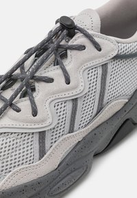 Scarpa sportiva grigio chiaro con tomaia in rete, dettagli in suede, design ondulato, lacci grigio scuro e suola in gomma screziata.