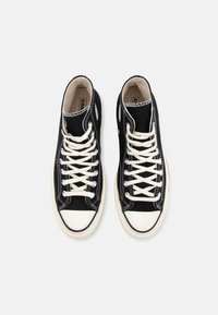 Sorte høyhælte sneakers laget av lerret, med hvite snørebånd, rund tåhette, kontrasterende sømmer og sidehull.