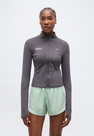 Femme aux cheveux tressés portant une veste Nike grise zippée et un short de sport Nike vert clair, debout devant un fond blanc uni.