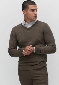 Brązowy sweter w serek wykonany z miękkiego materiału, z długimi rękawami i dopasowanym krojem, noszony na jasnej koszuli w paski.