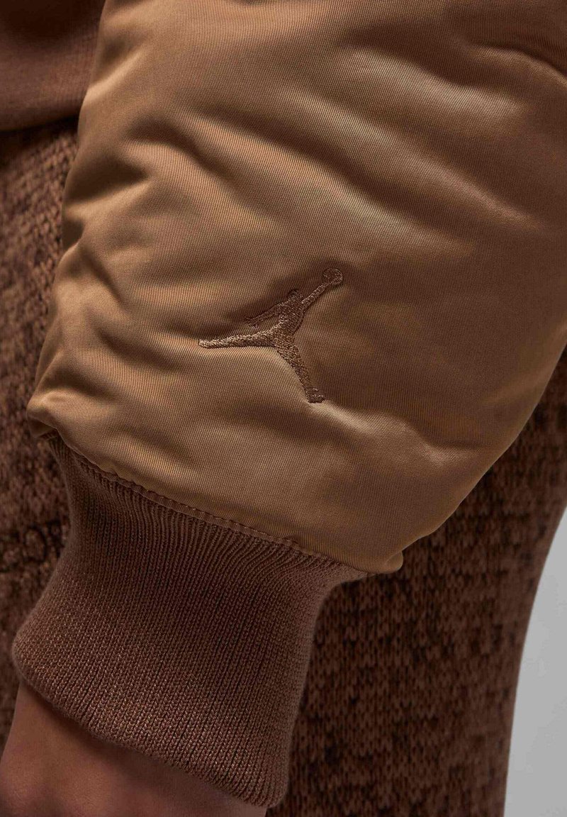 Sweatshirt en tissu beige avec une manche en maille texturée marron, arborant un logo Jumpman brodé ton sur ton sur la manche. Matière lisse avec des accents côtelés.