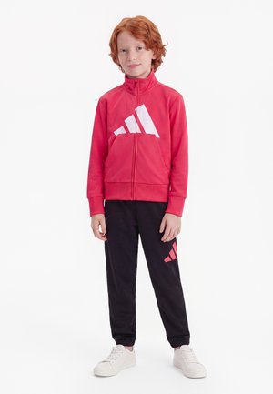 Enfant aux cheveux roux portant une veste rouge zippée et un pantalon noir avec des baskets blanches, debout sur un fond blanc.