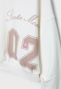 Sweat-shirt blanc avec le texte « Santa Monica », grand « 02 » en beige avec une petite étoile rose en haut à gauche du chiffre deux.