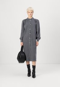 Grå denim skjortekjole med knappestolpe, rynkedetaljer på skuldrene, lange ermer og rett snitt, kombinert med en svart håndveske.
