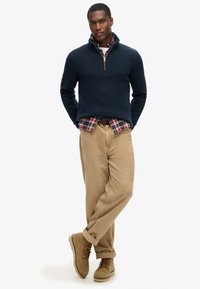 Marineblauer Strickpullover mit Reißverschlusskragen, über einem rot karierten Hemd getragen, kombiniert mit khakifarbenen Hosen und hellbraunen Schnürstiefeln.