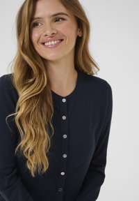Cardigan bleumarin dintr-un material moale, cu guler rotund și cinci nasturi metalici pe față, având mâneci lungi.