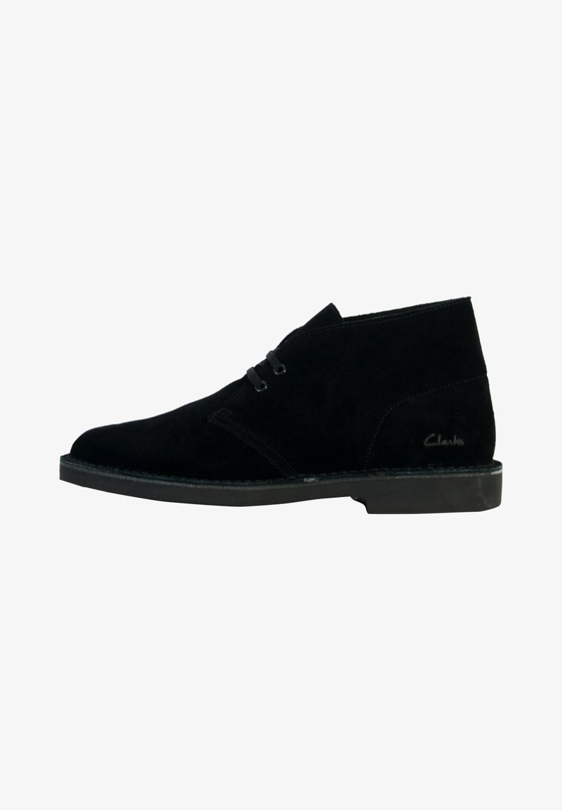 Stivaletti alla caviglia in suede nero con design chukka, dotati di due occhielli per i lacci e suola piatta in gomma. Nome del marchio "Clarks" visibile di lato.