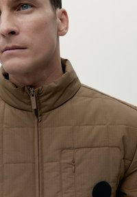 Giacca imbottita marrone con colletto alto, caratterizzata da una chiusura con zip, texture a quadretti e una piccola tasca sul petto con zip.