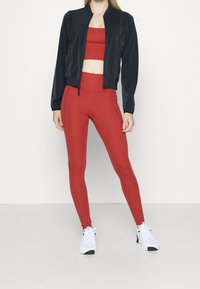 Chaqueta bomber negra con cremallera, sujetador deportivo rojo y leggings rojos de tiro alto; tejido suave, diseño ajustado, combinados con zapatillas deportivas blancas.