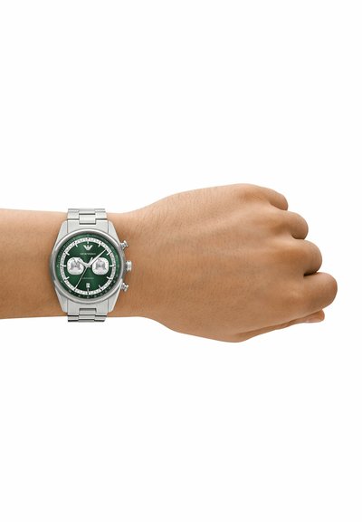 Poignet avec montre à bracelet métallique argenté, cadran vert, deux sous-cadrans argentés, tachymètre et affichage de la date à 6 heures.