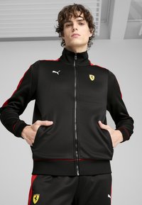 Zwarte trainingsjack met rode accenten, ritssluiting, hoge kraag en zijzakken; voorzien van het Puma-logo en het Ferrari-embleem op de borst.