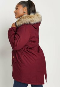 Burgundy parka dengan penutup kepala bulu tiruan, pinggang tali, penutupan ritsleting, dan saku samping. Tekstur lembut dengan desain ramping dan modern.