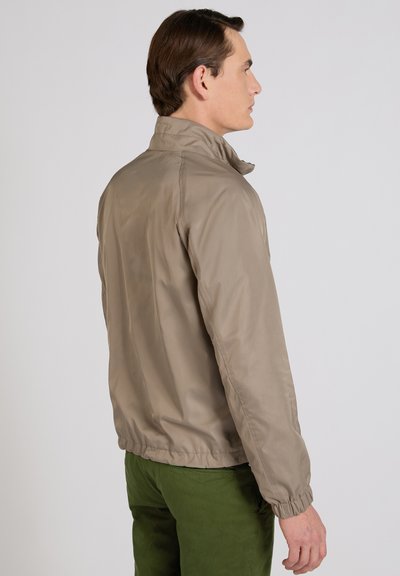 Uomo di profilo che indossa una giacca beige con polsini elasticizzati e pantaloni verdi su uno sfondo semplice grigio chiaro.