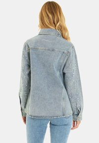Veste en denim bleu clair avec des manches ornées de rhinestones et de motifs cachemire. Col classique et poignets à boutons.