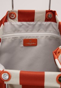 Interno di una borsa a righe rosse e bianche, con una tasca con zip e un'etichetta rettangolare in pelle arancione con branding in rilievo.