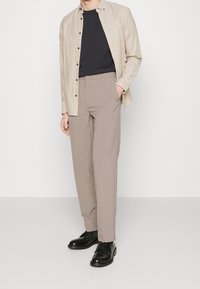 Béžová lněná košile, tmavé tričko s kulatým výstřihem, světle hnědé kalhoty a černé kožené boty. Volný střih, zapínání na knoflíky s našitými manžetami.