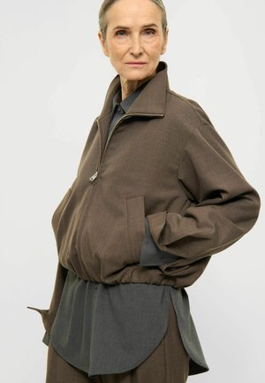 Femme plus âgée aux cheveux clairs portant une veste zippée marron par-dessus un t-shirt gris foncé, posant les mains dans les poches sur un fond uni.