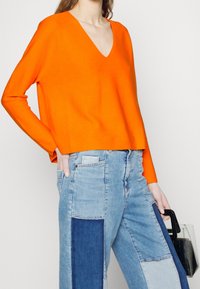 Oranges V-Ausschnitt-Pullover aus weichem Stoff, kombiniert mit einem Patchwork aus hellen und dunklen blauen Jeans, die einen lässigen Schnitt und Taschen haben.