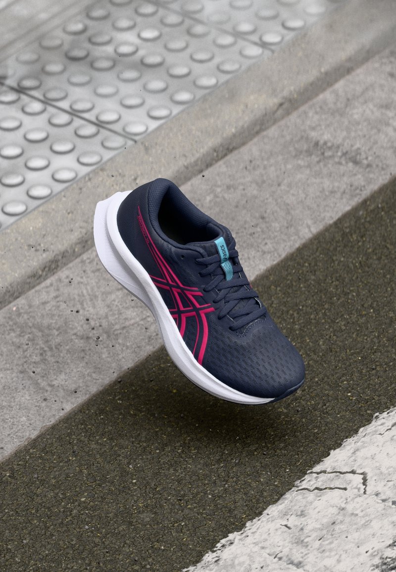 Chaussure de course bleu foncé et rose en l'air au-dessus d'un pavé texturé et du bord d'un trottoir carrelé.
