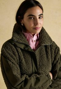 Oliwkowo-zielona kurtka polarowa z miękką, teksturowaną powierzchnią, wysokim kołnierzem i zapięciem na zamek z przodu. Noszona na jasnoróżowej koszuli z guzikami.