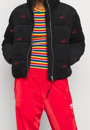 Person trägt eine schwarze gesteppte Jacke mit roten Logos, ein regenbogenfarben gestreiftes Hemd und leuchtend rote Adidas-Trainingshose, die Hände in den Taschen.