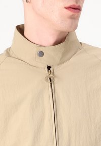 Beige lichtgewicht jacket met een opstaande kraag, rits aan de voorkant en metalen drukknoop. Heeft een gestructureerde stof en een ronde ritsstrop.