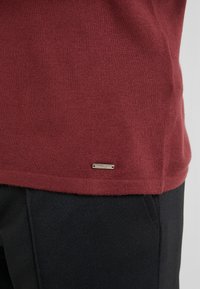 Pull bordeaux en tissu tricoté doux, avec une petite étiquette logo métallique à l'ourlet, associé à un pantalon foncé texturé.