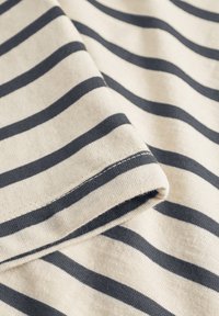 Közeli kép bézs színű szövetből, rajta egyenletesen elhelyezkedő vízszintes sötétkék csíkokkal és a hajtott szélén látható varrással.