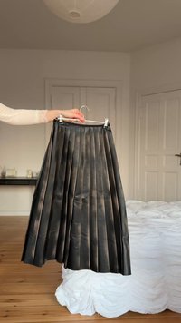 Χέρι κρατά πλισέ μαύρη δερμάτινη φούστα σε κρεμάστρα σε φωτεινό, μινιμαλιστικό υπνοδωμάτιο με λευκό κρεβάτι και ξύλινο πάτωμα.