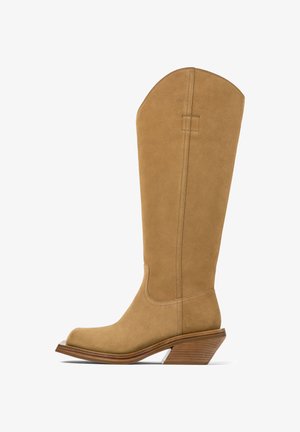 Beige suède laarzen tot aan de knie met een schuine houten blokhak en puntige neus, zichtbare stiksels langs de schacht en het voetgedeelte.