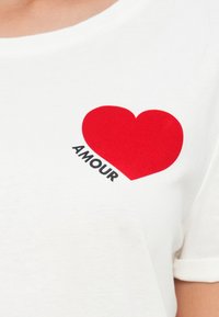 Λευκό T-shirt με γραφικό κόκκινης καρδιάς και τη λέξη "AMOUR" εκτυπωμένη διαγώνια κάτω από την καρδιά στο στήθος.