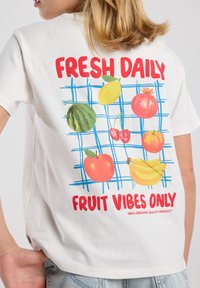 Camiseta de algodón blanca con gráficos coloridos de frutas, incluyendo sandía, cerezas y manzanas, con el texto "FRESCO A DIARIO" y "SOLO VIBRAS FRUTALES."