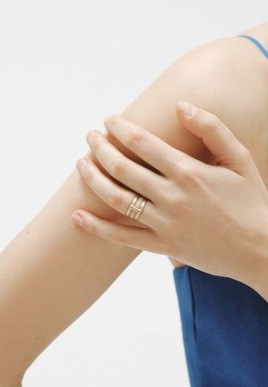 Mano con anillo dorado de doble banda descansa sobre el brazo superior, persona vestida con prenda azul sin mangas.