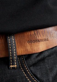 Brauner Ledergürtel mit eingeprägtem "DSQUARED2", durch dunkle Jeans mit sichtbarer Naht gefädelt, kombiniert mit schwarzem Stoffoberteil.