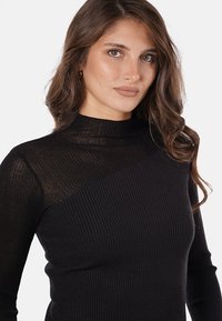 Maglione nero a collo alto con costine, arricchito da un pannello trasparente su una spalla, che evidenzia la texture e gli elementi di design a contrasto.