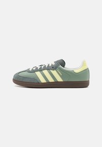 Green and grey suede sneaker with three yellow stripes, white laces, and a textured brown rubber sole. Features a padded collar and tongue.