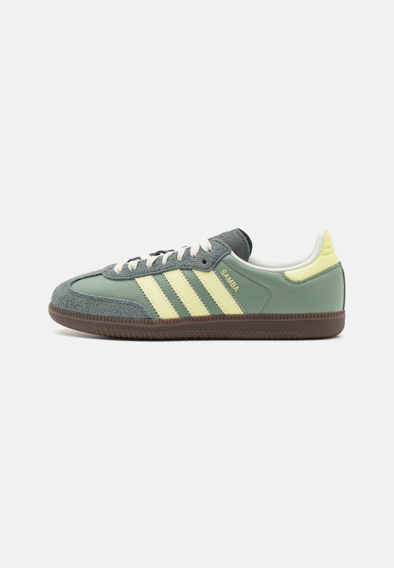 Green and grey suede sneaker with three yellow stripes, white laces, and a textured brown rubber sole. Features a padded collar and tongue.