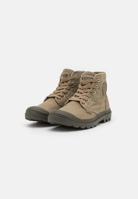 Botas de tobillo de lona beige con suelas de goma oscura, puntera redonda, ojales de metal y parche con logo en el lateral. Cuentan con cordones para el cierre.