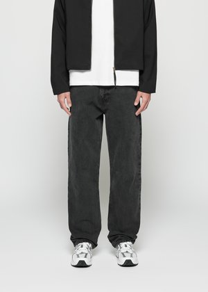 Man wearing black jacket, white t-shirt, loose dark jeans, and white sneakers standing against plain background.