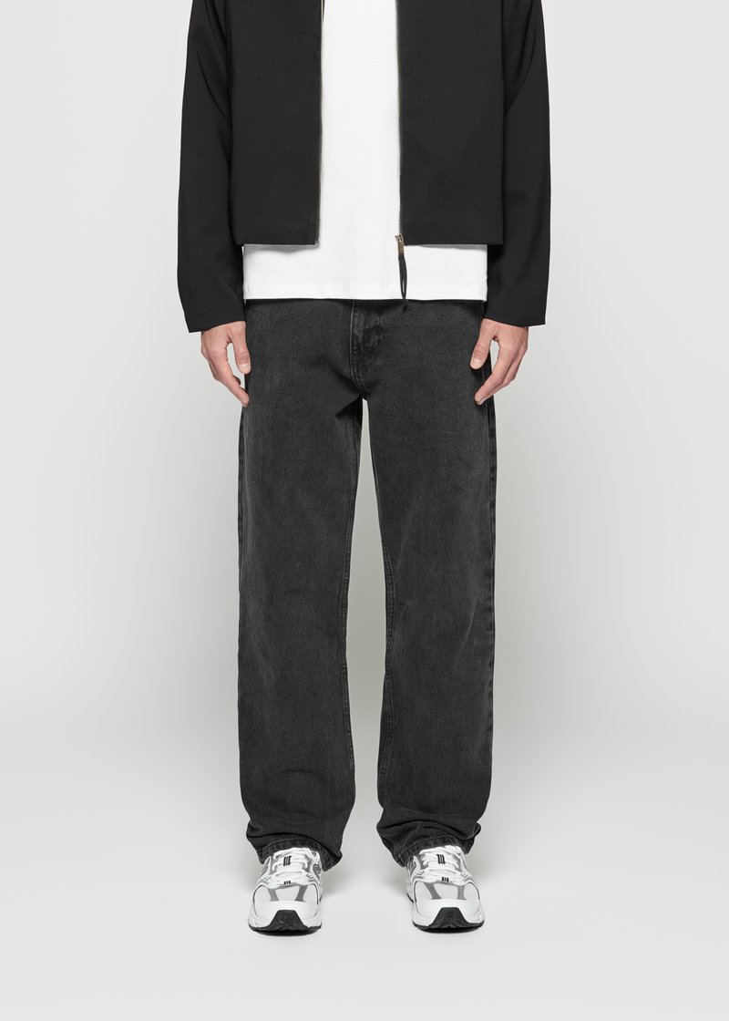Mann med svart jakke, hvit t-skjorte, løse mørke jeans og hvite joggesko som står mot en enkel bakgrunn.