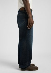 Dark blue denim jeans with a relaxed fit, featuring a leather Lee brand patch, five-pocket design, and visible stitching. Brown footwear is worn.