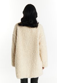 Palton din blană de shearling cream, moale, cu o croială lejeră, mâneci lungi și guler rotund. Prezintă un material texturat și o croială de lungime medie.